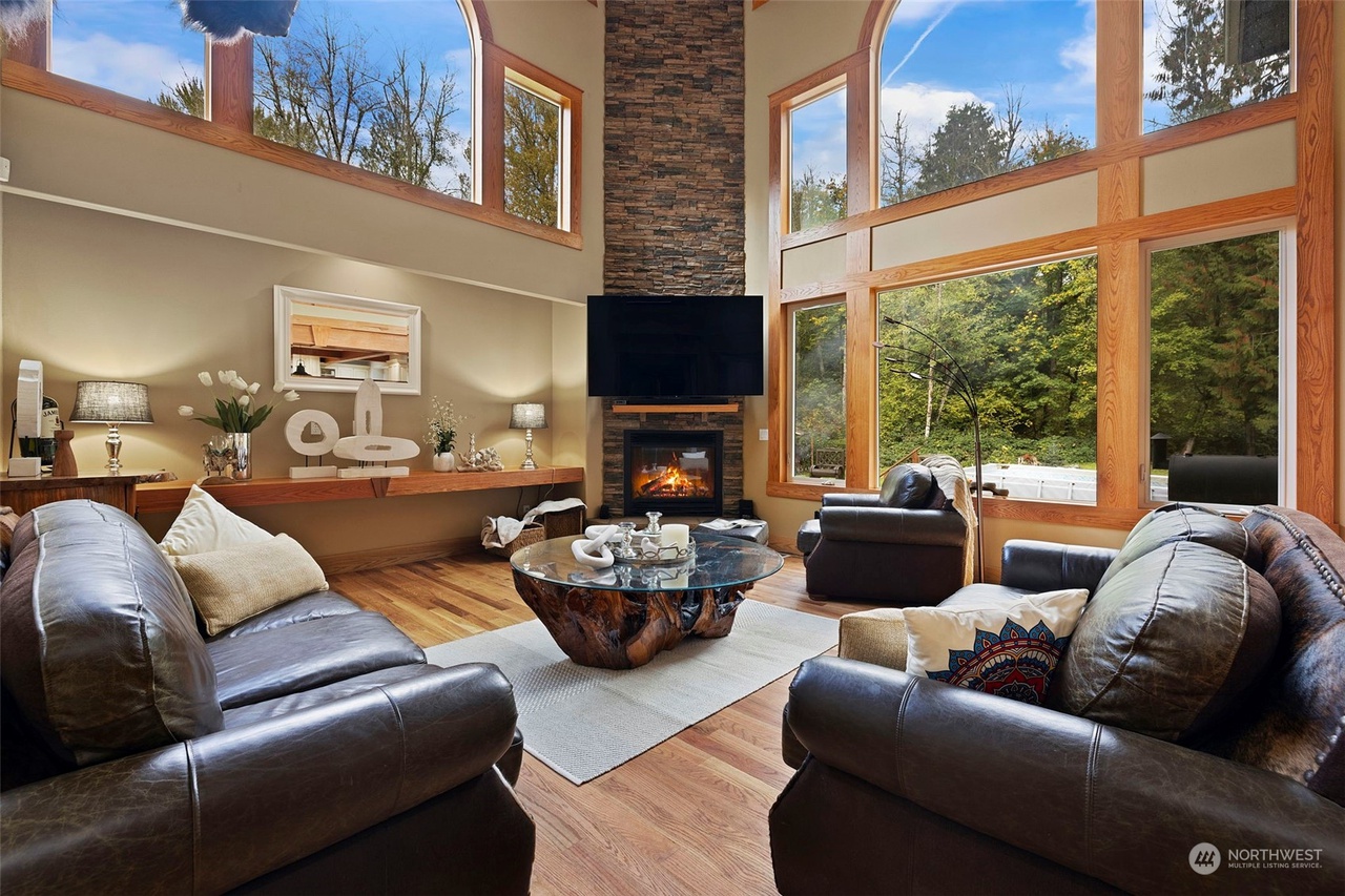 Spacious living room with large windows, leather sofas, a stone fireplace, and a glass coffee table. Forest view outside, perfect for Seattle's nature-loving home stagers.